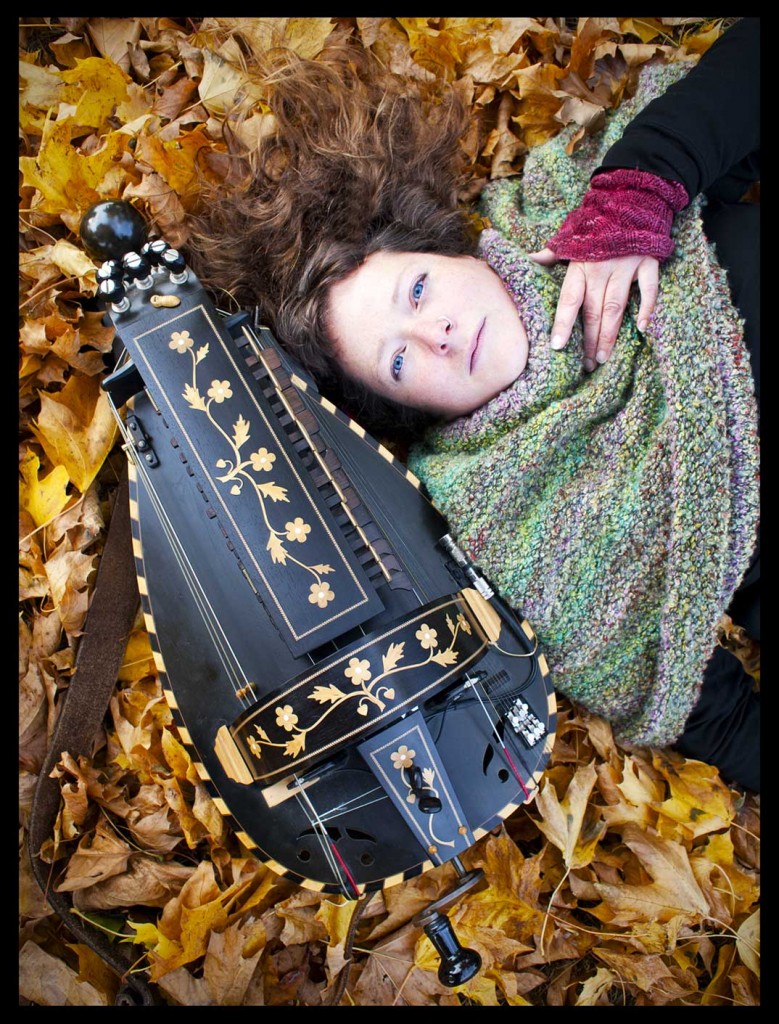 Jess lying on a bed of autumn leaves next to a black hurdy gurdy with natural wood inlay of flowers