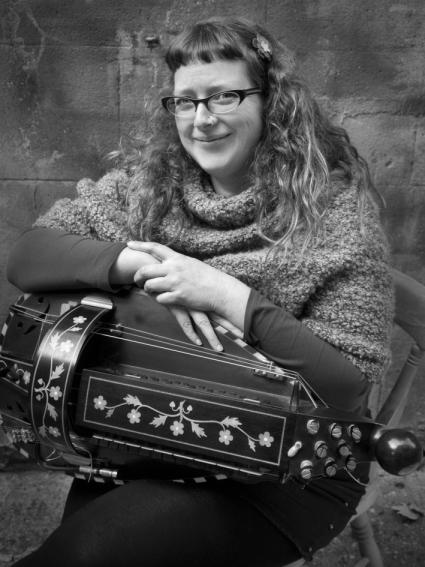 A black and white image of Jess seated, holding a black hurdy gurdy with natural wood coloured decoration. She has long brown hair and glasses and is smiling